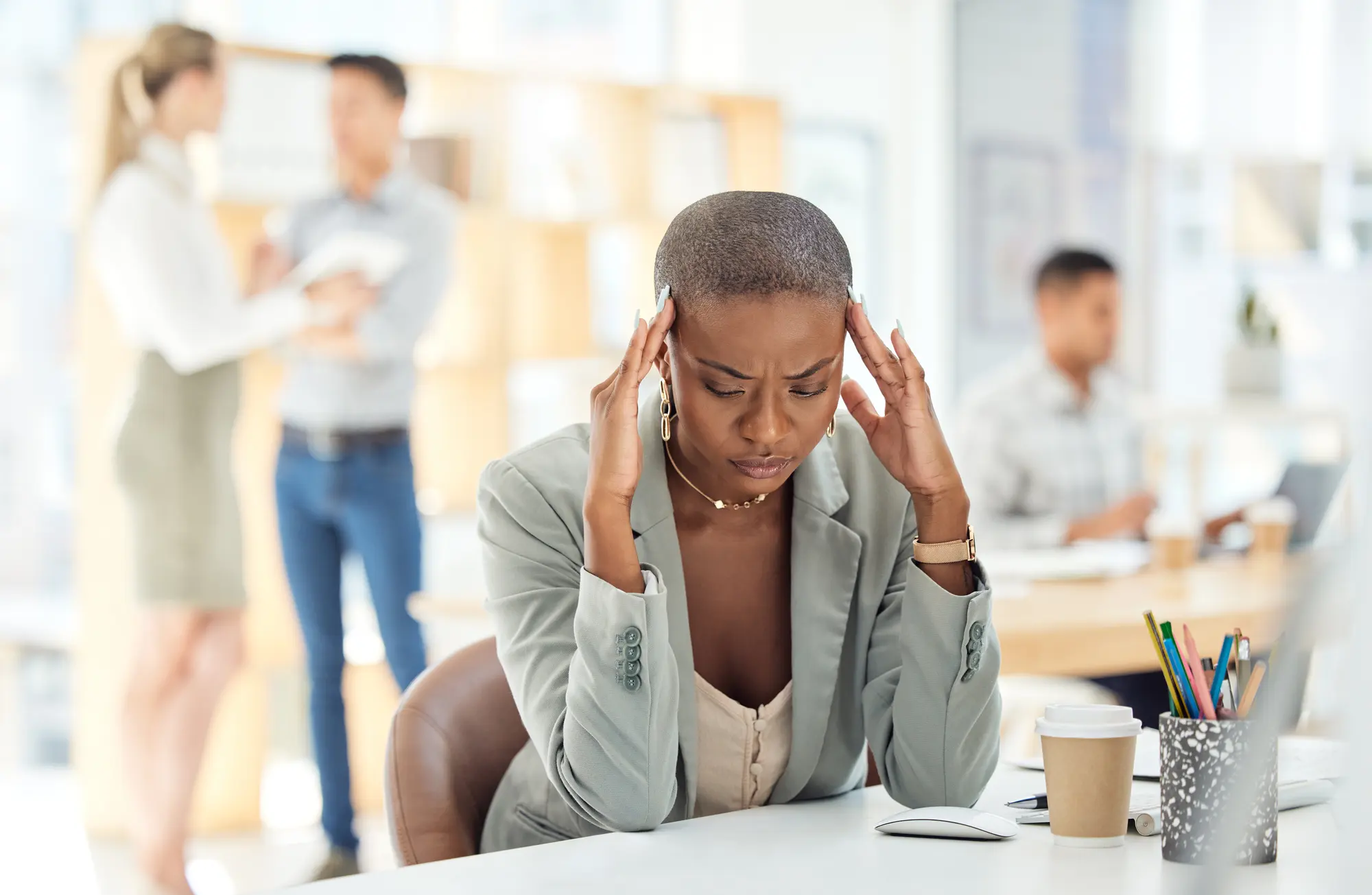 Una donna in ufficio si tiene le tempie, stanca, mentre i colleghi parlano sullo sfondo